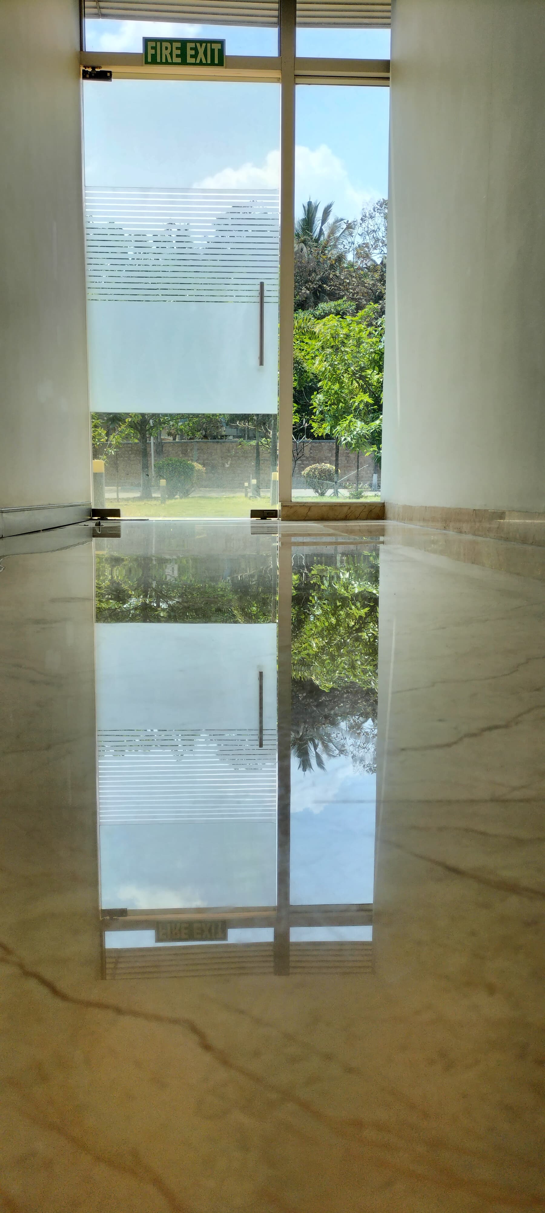 Polished marble floor perfectly reflecting a glass fire exit door and lush green trees.